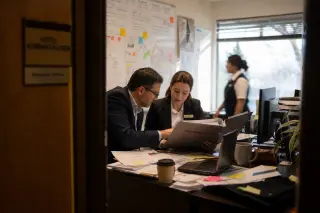 Two hotel managers reviewing insurance documents in a naturally lit, slightly cluttered office with a staff member walking by in the background, captured in the midst of real work.