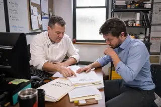 Two managers in a restaurant back office reviewing insurance papers together in a focused discussion, natural light coming through window, candid scene.