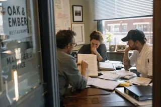 Restaurant office scene with three staff members intently reviewing documents at a cluttered desk, natural light streaming through blinds, conveying a candid moment of focused discussion.