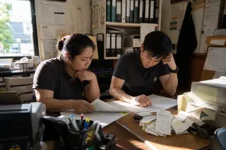 Two restaurant managers reviewing complicated paperwork in a cluttered office, focused and concerned, natural light creating a serious and authentic workspace mood
