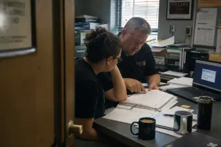 Two restaurant managers focused on paperwork and discussion in a cluttered back-office, natural light creating a real and serious atmosphere.